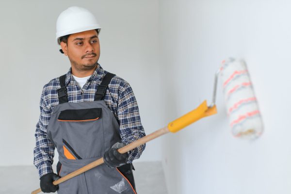 An Indian apartment repair worker paints a white wall with a roller