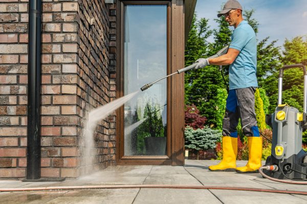 A person is cleaning a brick wall with a pressure washer outside a house while standing on a patio.